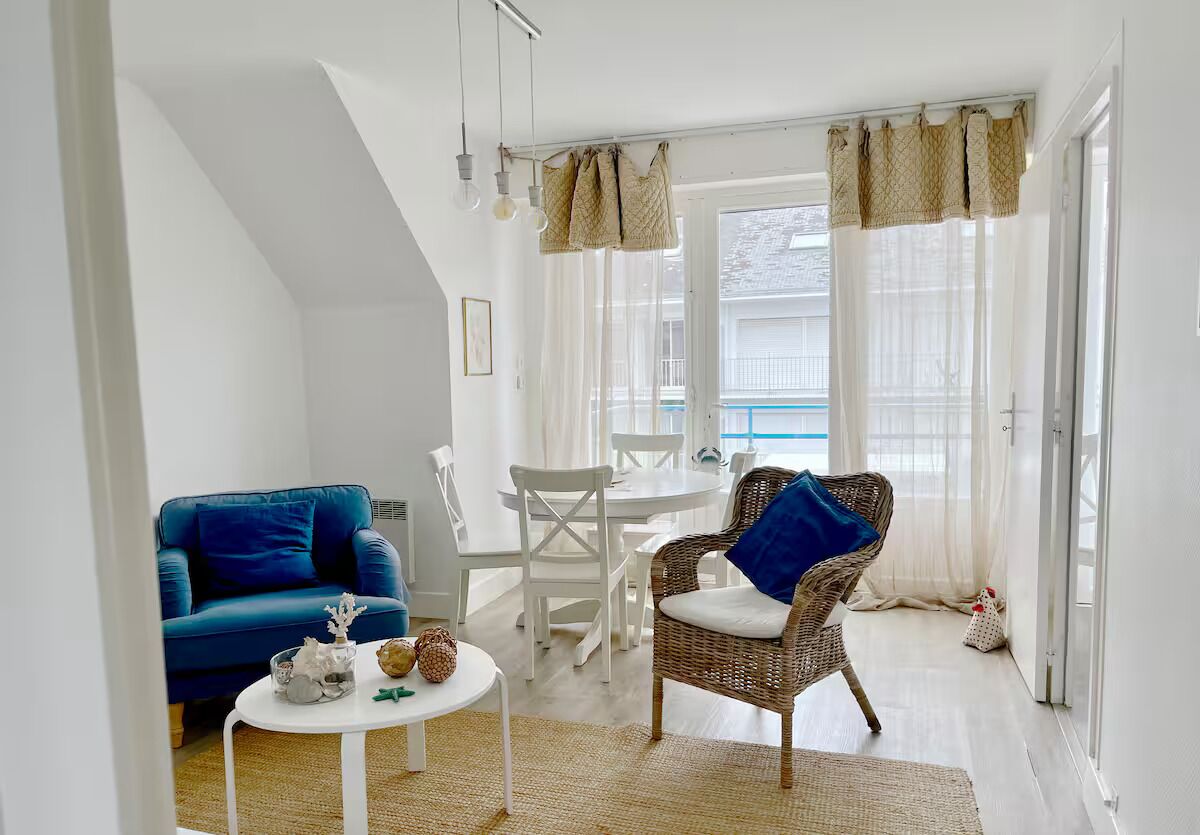 Un salon chaleureux avec un canapé bleu, un fauteuil en osier, une petite table ronde et une table à manger avec des chaises.