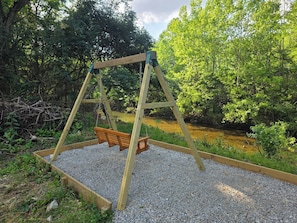 Un ensemble de balançoires en bois installé dans un jardin dont le sol est recouvert de gravier, entouré de verdure et bordé par un ruisseau.