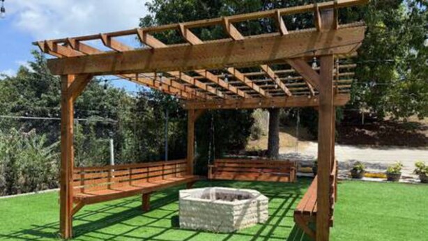 A wooden pergola structure with a stone fire pit in the center, surrounded by a well-maintained lawn. - Oasis at Hacienda el Lago (Cayey)