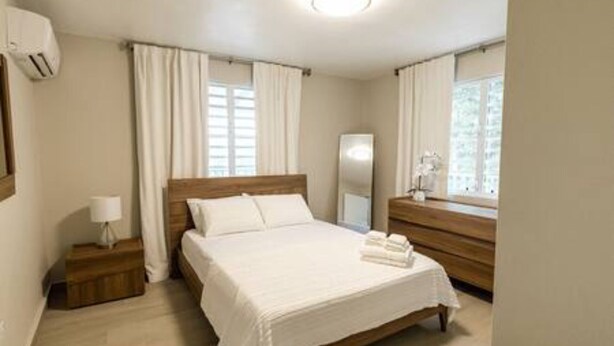 A bedroom with a wooden bed, white linens, a bedside table with a lamp, and a window with curtains. - Serenidad at Hacienda el Lago (Cayey)