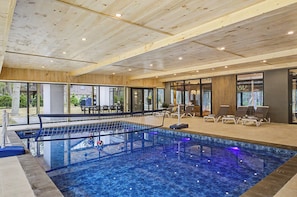 An indoor swimming pool with a wooden ceiling, lounge chairs, and a view of the outdoors. - New Modern Creekfront Mansion with Indoor Pool and Sauna by Camelback (Tannersville)