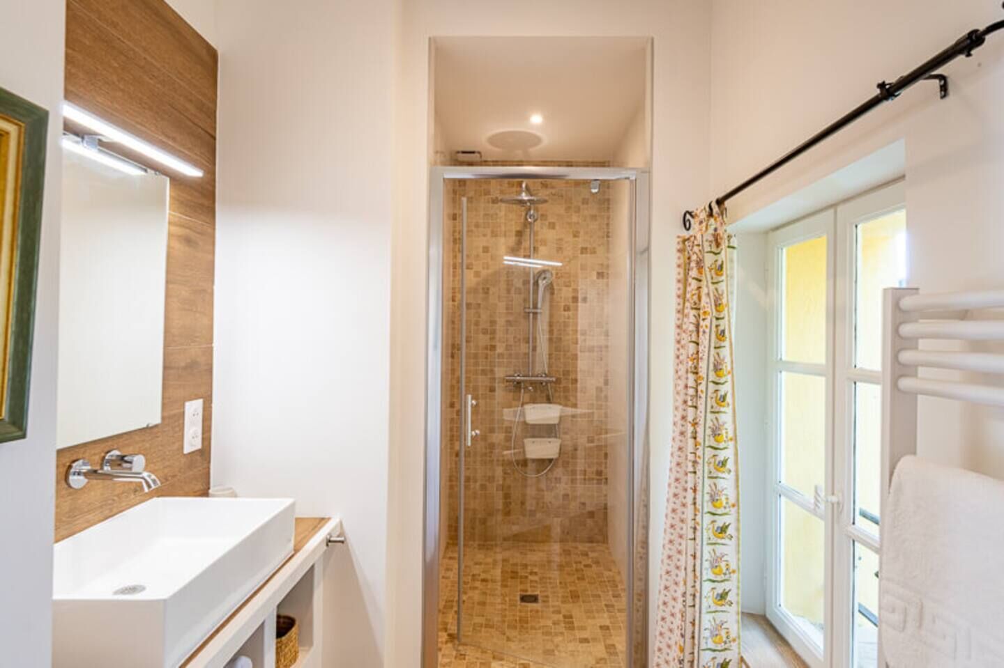 Une salle de bain avec une douche en verre, un lavabo, un miroir et une fenêtre avec un porte-serviettes.