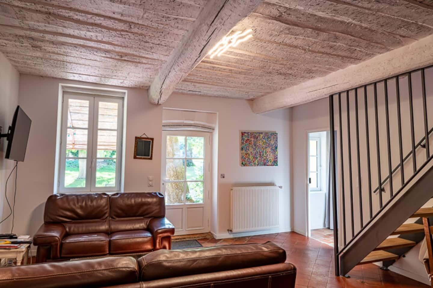 Un salon avec un canapé en cuir marron, un téléviseur, une fenêtre, une porte, un radiateur et un escalier.