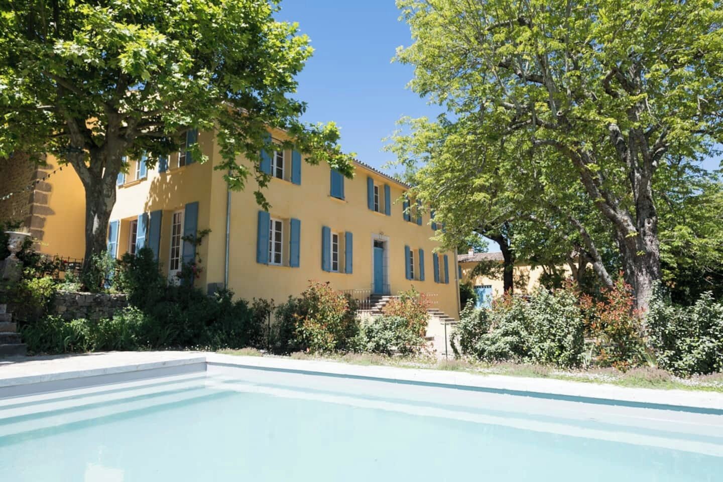 Un bâtiment jaune avec des volets bleus, une piscine et de la verdure tout autour.