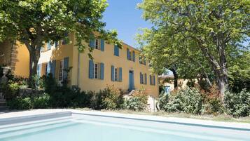 Un bâtiment jaune avec des volets bleus, une piscine et de la verdure tout autour.