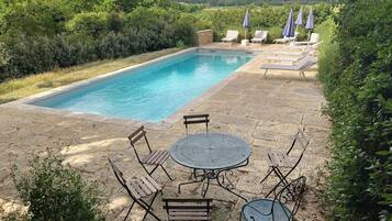 A rectangular pool with a stone deck, surrounded by lounge chairs and umbrellas, set in a lush garden.