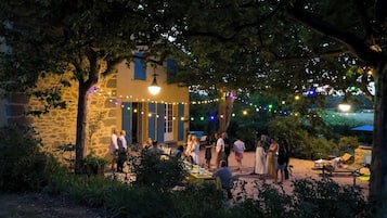 An outdoor gathering with people seated at tables under trees, a stone building with blue shutters, and string lights in the background.