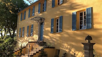 Un bâtiment à deux étages, doté d’une façade jaune, de volets bleus, d’un escalier menant à une entrée abritée et d’un pilier en pierre orné d’une plante.