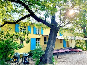 A yellow building with blue shutters, a large tree in front, and outdoor seating. - Château de Trians – 3 Pools, 19 Bedrooms, Sleeps up to 48 (Néoules)