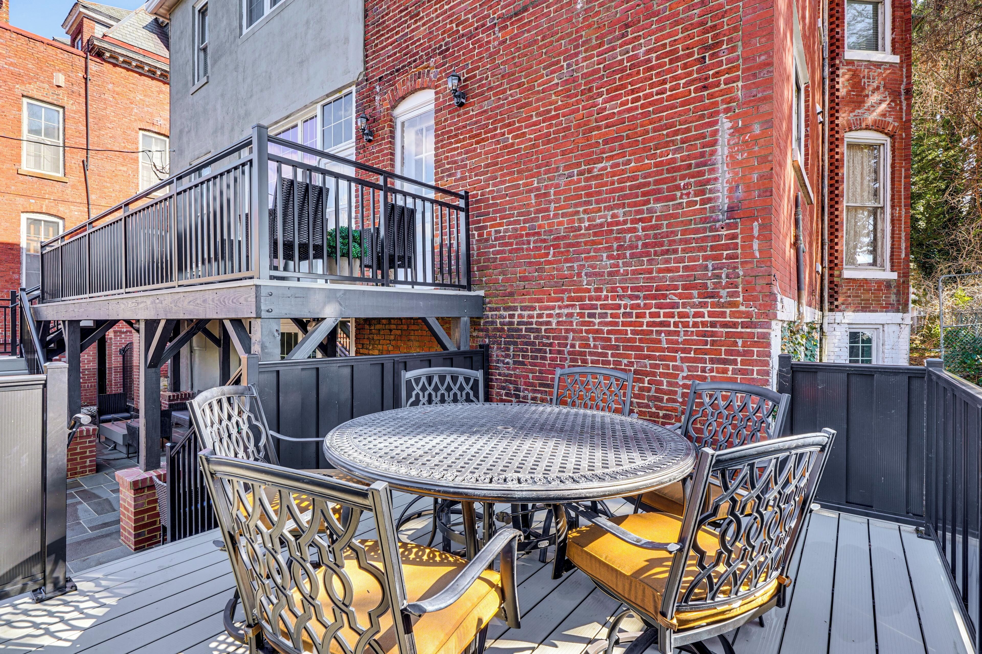 A deck with a round table and chairs, a small outdoor kitchen, and a brick building in the background.