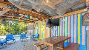 A wooden deck with a table and chairs, a TV mounted on the wall, and a colorful striped wall. - Sun & Sand in St Pete! Family Home w/ Tiki Bar (St. Petersburg)