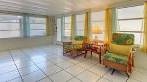 A room with a tiled floor, two wicker chairs with floral upholstery, a small wooden table with a lamp, and a window with yellow curtains. - Sun & Sand in St Pete! Family Home w/ Tiki Bar (St. Petersburg)