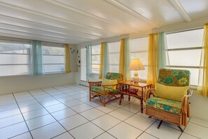 Ein Zimmer mit Fliesenboden, zwei Korbstühle mit floralem Bezug, ein kleiner Holztisch mit Lampe und ein Fenster mit gelben Vorhängen.