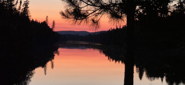A calm lake reflecting trees and a mountain range at sunset.