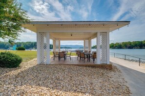 Een overdekt terras met tafels en stoelen, met uitzicht op een waterpartij en bergen.
