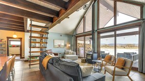 Amplia sala de estar con un sofá grande, una mesa de comedor y una chimenea. Varias ventanas ofrecen vistas al exterior.