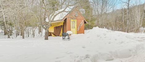 A wooden cabin with a yellow door, surrounded by snow-covered ground and leafless trees.