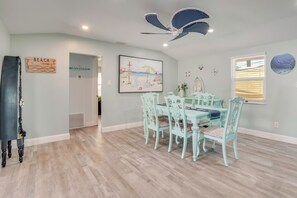 Un comedor con mesa y sillas, ventilador de techo y adornos de pared que incluyen una escena de playa y un letrero.