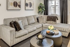 Een moderne woonkamer met een grijze hoekbank, een ronde salontafel met decoratieve objecten, en kunstwerken aan de muren.