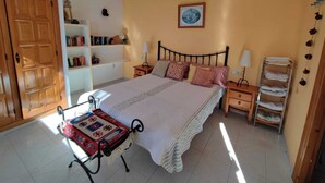 A bedroom with a bed, bedside tables, a nightstand, a shelf with books, a hanging lamp, and a door.