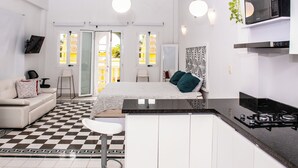 A modern hotel room with a checkered floor, a bed with green pillows, a white sofa, a small white chair, a kitchen area with a stove, and a TV mounted on the wall. - Charming Studio with Balcony + Pool on Santo Domingo square, old city (Cartagena de Indias)