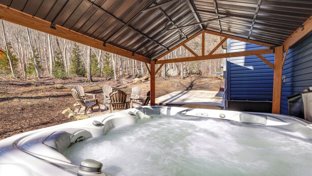 A hot tub under a metal roof with wooden chairs and a view of trees. - Lake Lure Getaway w/ Hot Tub & Fire Pit! (Lake Lure)
