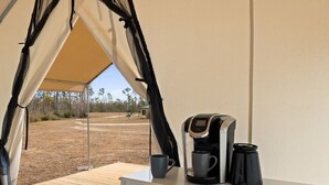 A tent with a coffee maker and two mugs on a wooden deck, overlooking a dry landscape with sparse vegetation and a clear sky.