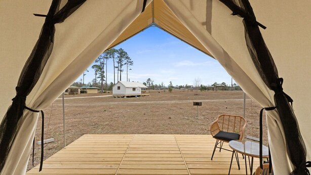 A tented area with a wooden deck, two wicker chairs, and a view of a small house in the distance.