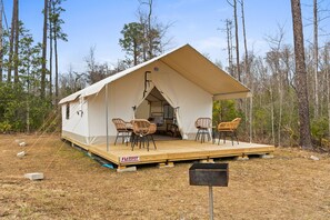 Un camping-car à plateforme, doté d’un porche couvert et de quatre chaises en osier, installé dans une zone boisée.