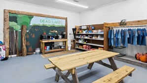 Una habitación con un mural que dice 'Welcome to Dead Lakes', una mesa de madera, estantes con diversos objetos y ropa colgada.