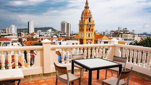 A terrace with a table and chairs, overlooking a cityscape with a prominent yellow building. - Charming Colonial Studio with Balcony on Santo Domingo Square + Pool (Cartagena de Indias)