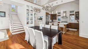 A spacious dining area with a dark wooden table and white chairs, a staircase leading to an upper level, and a modern kitchen with stainless steel appliances. - Private Beach Access | Bikes | Patio (Watersound)