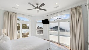 Une chambre à coucher avec un lit, un ventilateur de plafond, une télévision et de grandes fenêtres avec des rideaux.