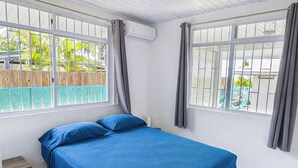 Uma cama de solteiro com roupa de cama azul num quarto com paredes brancas e janelas amplas.
