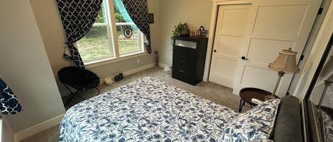 A bedroom with a bed, a dresser, a desk, a chair, a window with curtains, a ceiling fan, and a door.