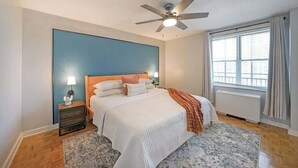 Une chambre à coucher comprenant un lit, des tables de chevet, un ventilateur de plafond, une fenêtre avec des rideaux et un radiateur.