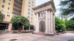 Une place publique avec un monument de style classique, un bâtiment portant une enseigne indiquant « TOWERS », et un arbre.