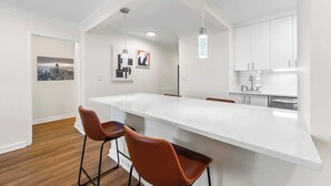 Una cocina moderna con armarios blancos, una isla grande con taburetes y luces colgantes, además de vistas a la ciudad a través de un ventanal.