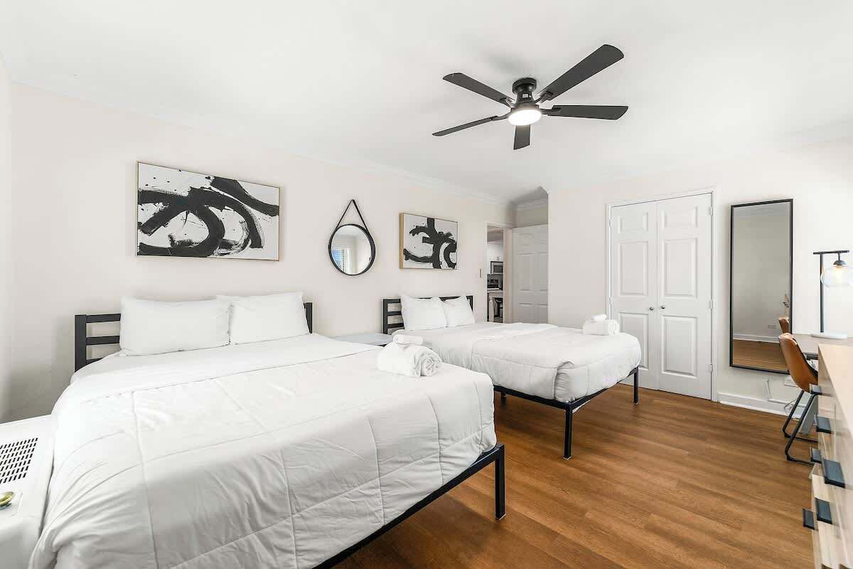 A hotel room with two beds, a ceiling fan, artwork on the walls, a desk with a chair, and a door leading to another room.