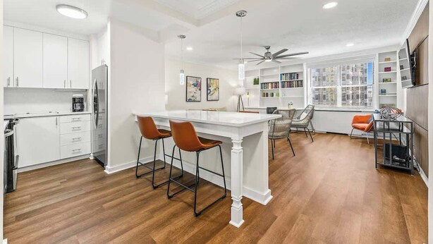 Una cocina moderna con armarios blancos, una isla central con taburetes de bar, un ventilador de techo y vistas a una biblioteca con estanterías.