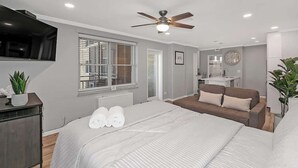 Ein modernes Schlafzimmer mit einem großen Bett, einem Deckenventilator, einem Fernseher und einem Fenster mit Jalousien.