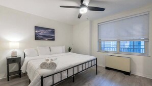A bedroom with a bed, a ceiling fan, a window with blinds, a radiator, and a nightstand with a lamp. - Bright Downtown Retreat w/ Full Kitchen (Atlanta)