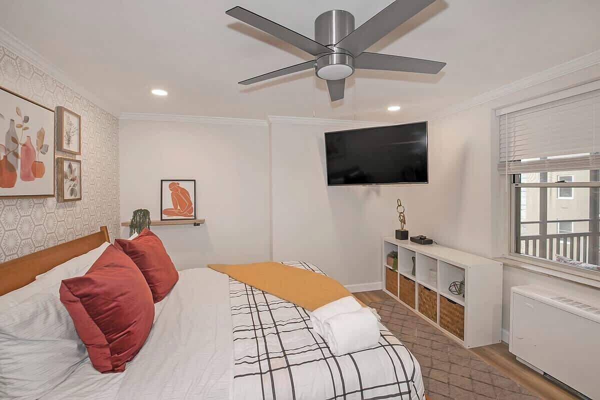 A bedroom with a bed, a ceiling fan, a TV, and a window with blinds.