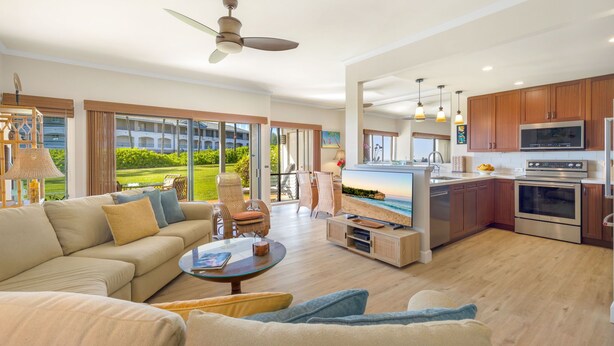 A spacious living room with a large sofa, a coffee table, and a television. There is a kitchen area with modern appliances and wooden cabinets. The room has large windows with a view of a green landscape. - VERY SPECIAL  Renovated 1 Bdrm Condo - A/C - Oceanview, Athletic Club Access (Koloa)