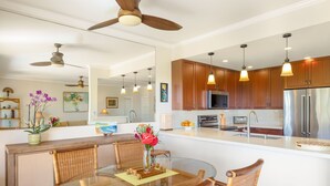 A modern kitchen with wooden cabinets, stainless steel appliances, and a glass dining table with wicker chairs. - VERY SPECIAL  Renovated 1 Bdrm Condo - A/C - Oceanview, Athletic Club Access (Koloa)