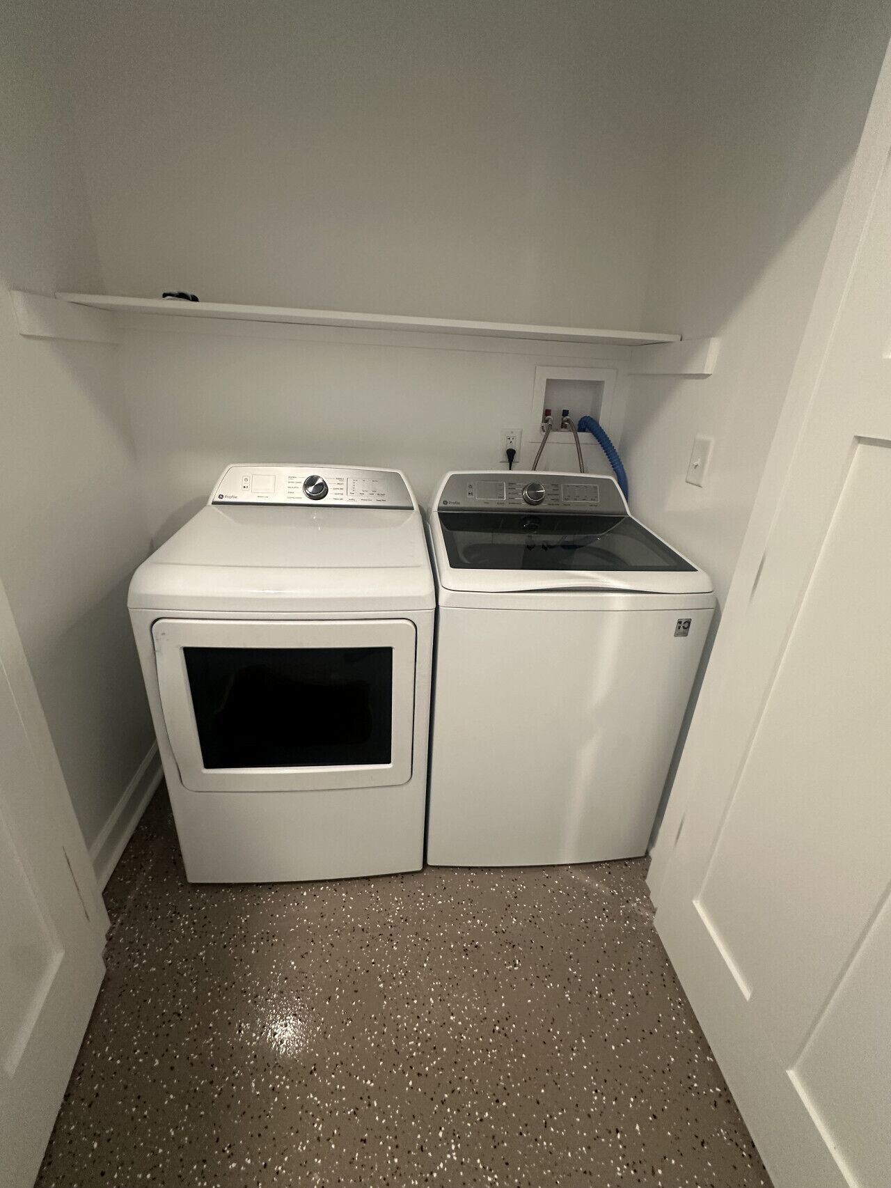 A laundry room with two washing machines and a dryer, a white shelf above, and a speckled floor.