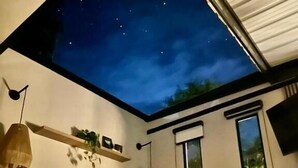 Une chambre moderne avec une verrière au plafond, un lit avec une literie blanche, une étagère fixée au mur et une grande fenêtre.