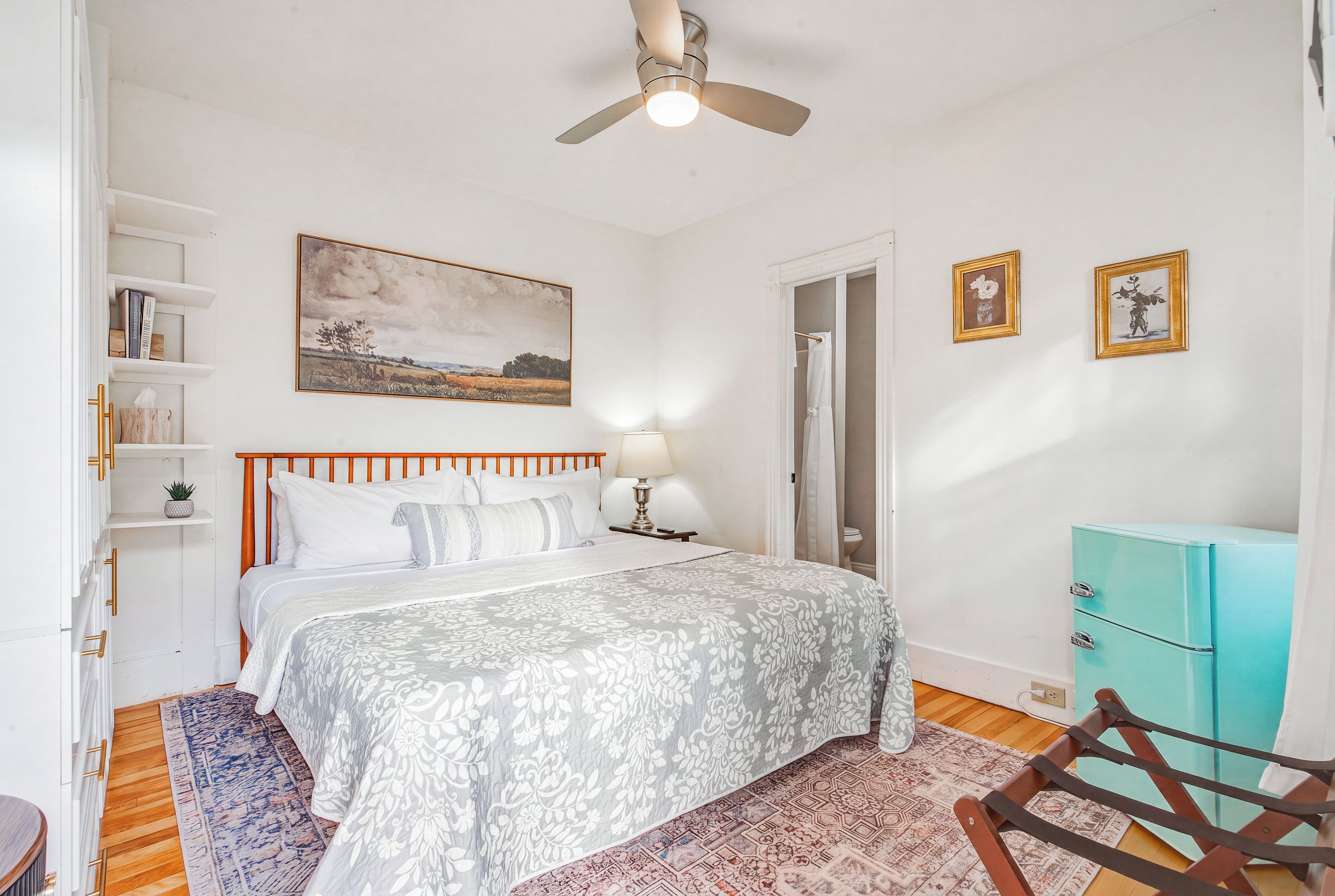 A bedroom with a bed, a ceiling fan, a nightstand, a lamp, a dresser, a chair, and a door leading to a bathroom.