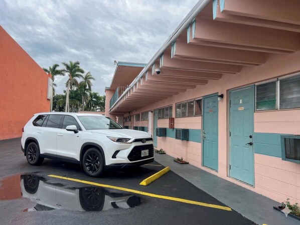 A white SUV parked in a building with a pink and teal color scheme, a blue door labeled '3', and a small garden area with plants. - Anhinga Motel Homestead Fl (Homestead)