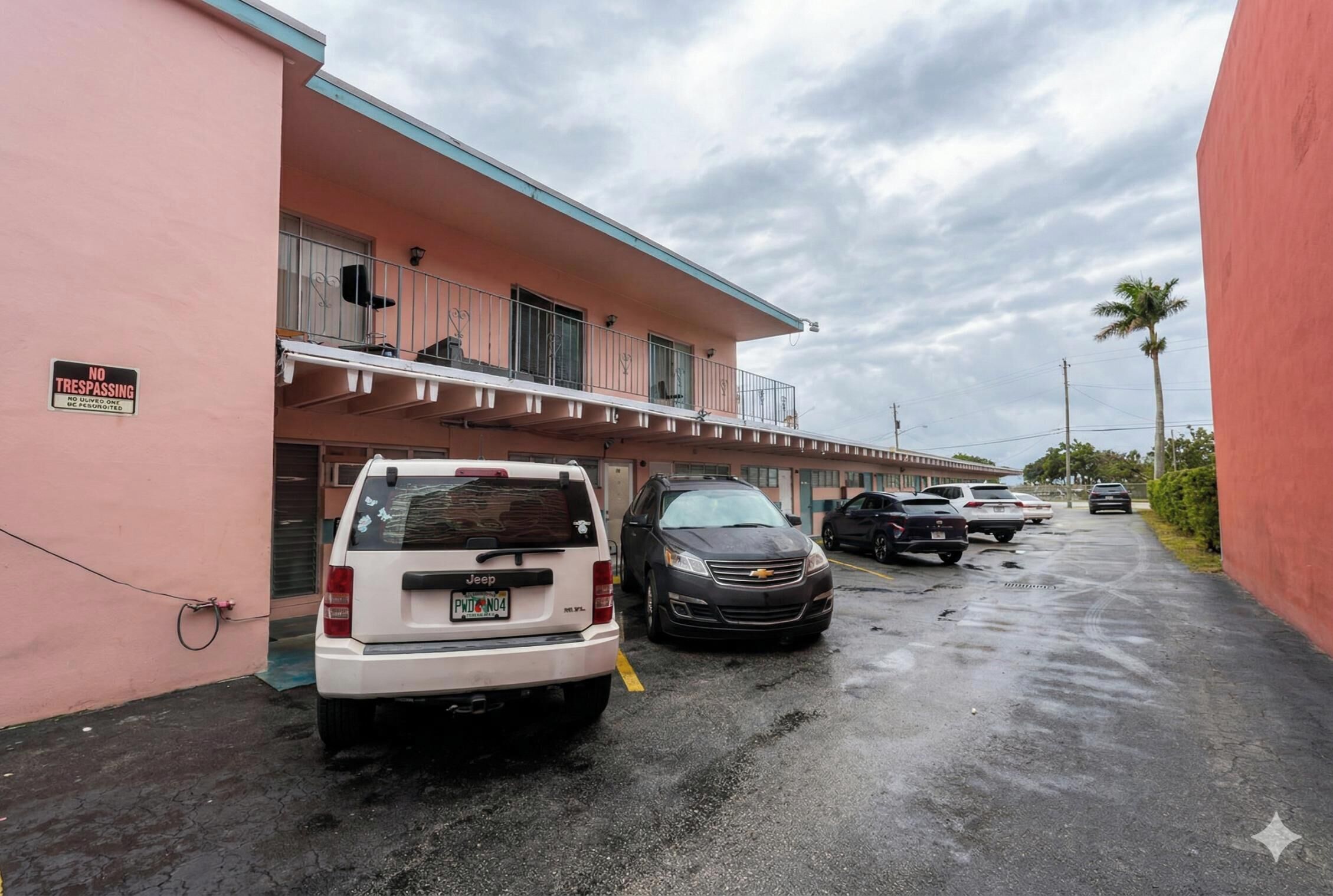 A parking area with a white Jeep and a black car parked in front of a pink building with a 'NO TRESPASSING' sign.